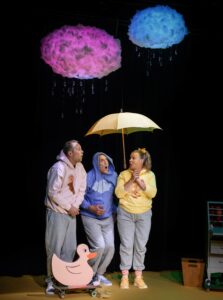 Tre personer i pastelliga kläder står under ett ljusgult paraply på en mörk scen. Ovanför dem regnar det från två fluffiga moln.