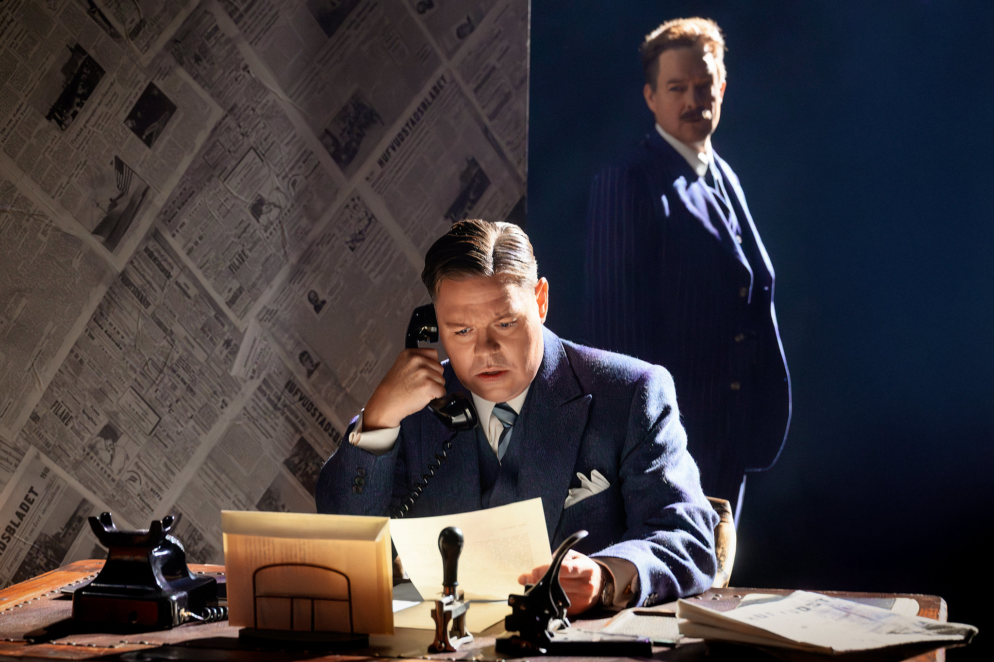 En man i blå kostym sitter vid ett skrivbord och pratar i telefon samtidigt som han läser ett dokument. Bakom honom står en annan man i mörk randig kostym, delvis dold i skuggorna.