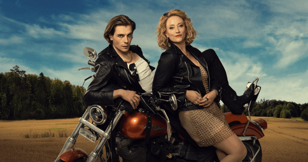 Mikko Kauppila och Sannah Nedergård som Zac och Fanny i musikalen Änglagård. De poserar framför en orange motorcykel på en landsvägsliknande plats. Båda bär svarta läderjackor, och Fanny sitter på motorcykeln i en ljus klänning med dekorativa detaljer. I bakgrunden syns åkerlandskap och blå himmel.