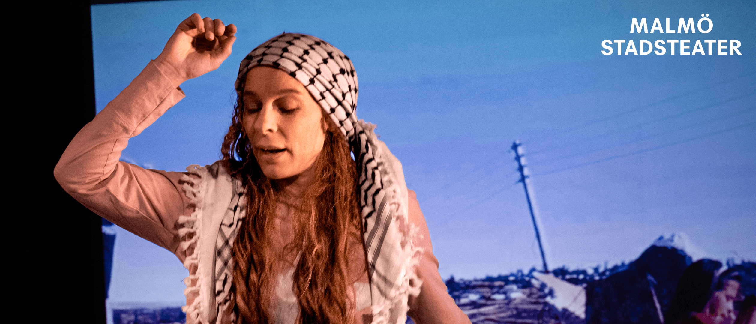 En person står på en scen med en svartvit palestinasjal över håret och en rosa långärmad tröja. Bakom syns en projicerad bakgrund med ett landskap av sten och elstolpar.