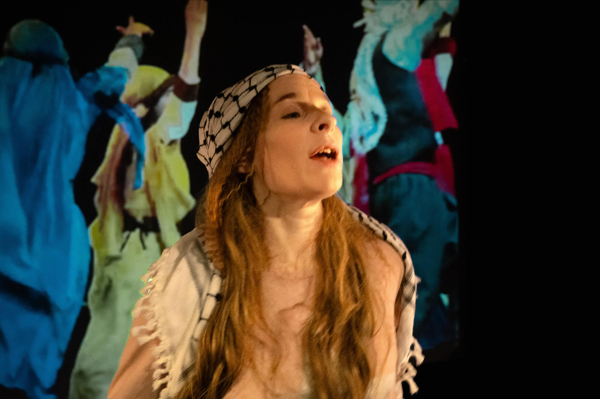 En person står på en scen med en svartvit palestinasjal över håret och en rosa långärmad tröja. Bakom syns en projicerad bakgrund med ett landskap av sten och elstolpar.