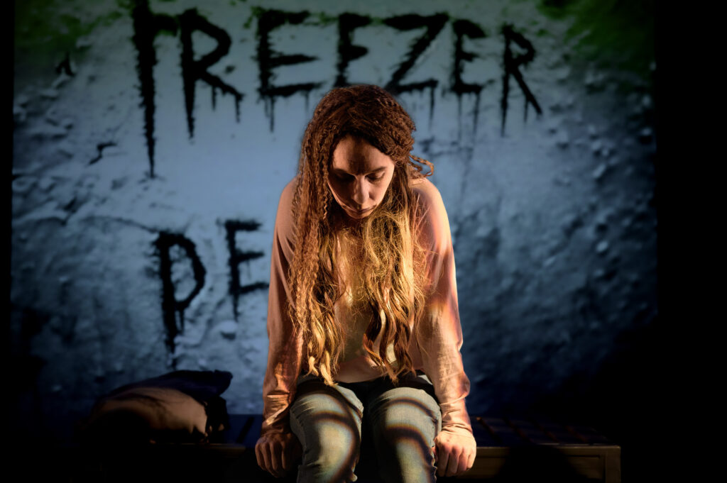 En person sitter på en bänk i ett dunkelt belyst rum med en mörk bakgrund. På väggen bakom syns stora, svarta bokstäver som bildar orden ”FREEZER”