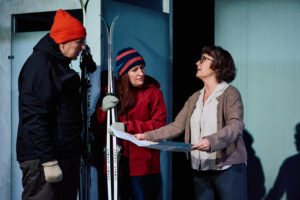 Två personer klädda i vinterjackor och mössor står med skidor lutade mot väggen, medan en tredje person håller ett stort papper eller karta. Scenen har kalla färger och betongliknande väggar. Det är skådespelarna Mitja Sirén, Nina palmgren och Anna Hultin.