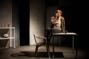 Skådespelaren Anna Hultin sitter på kanten av ett bord i ett sparsamt möblerat rum med grå väggar. Framför står en plaststol, och på bordet ligger en kopp. Till vänster syns en liten hylla med några föremål, bland annat en cykelhjälm och en flaska. Ljuset är varmt och riktat mot personen, vilket skapar starka skuggor i rummet.