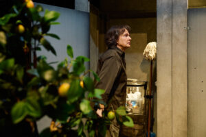 Skådespelaren Anna Hultin står vid en städvagn med rengöringsmedel och en mopp, placerad i en smal öppning mellan grå väggar. Till vänster syns en grön växt med gula frukter som ger kontrast mot den industriella miljön.