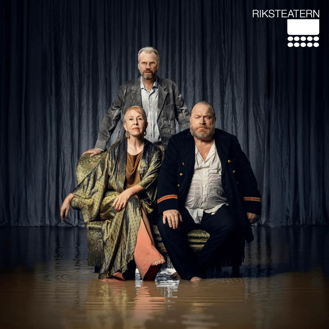 Tre personer sitter och står framför en mörk, draperad bakgrund. En kvinna sitter i mitten i en mönstrad klänning, en man i mörk kavaj sitter bredvid henne, och en tredje man står bakom dem. Alla ser allvarliga ut.