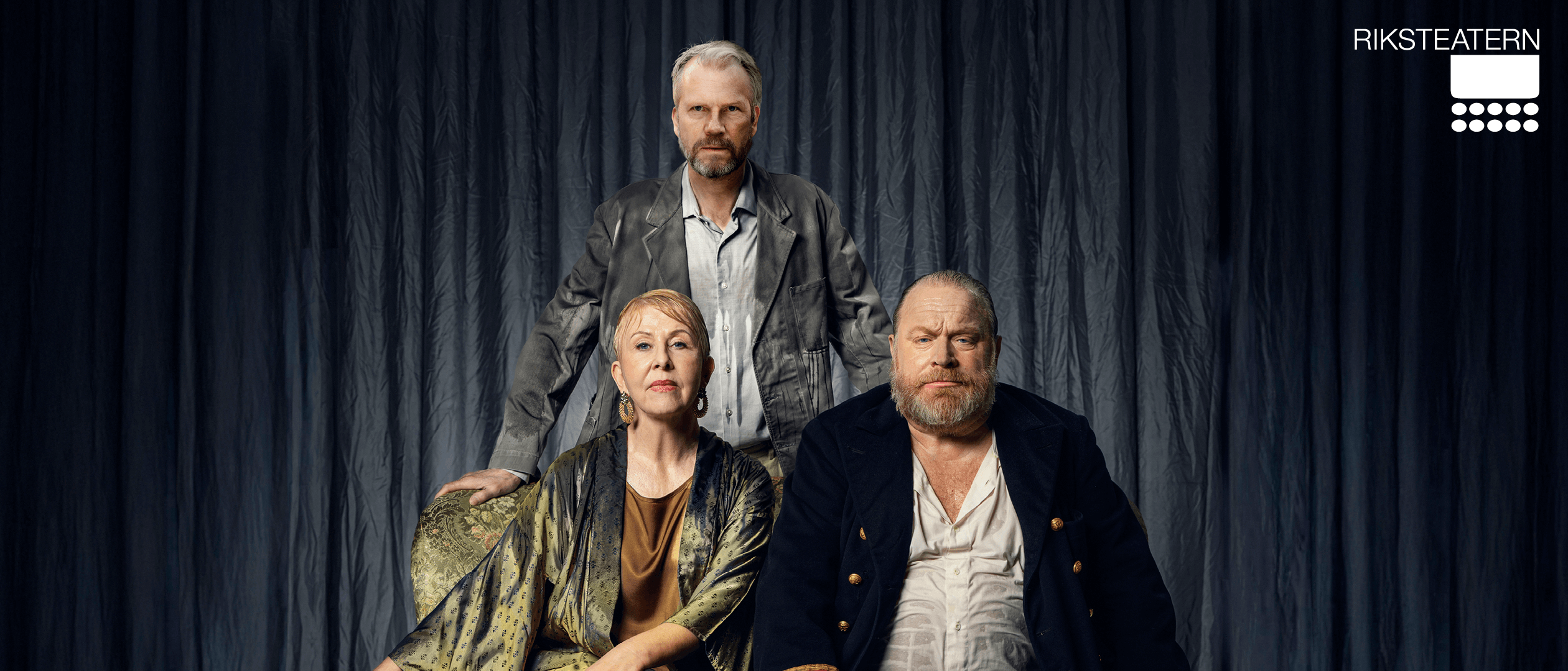 Tre personer sitter och står framför en mörk, draperad bakgrund. En kvinna sitter i mitten i en mönstrad klänning, en man i mörk kavaj sitter bredvid henne, och en tredje man står bakom dem. Alla ser allvarliga ut.