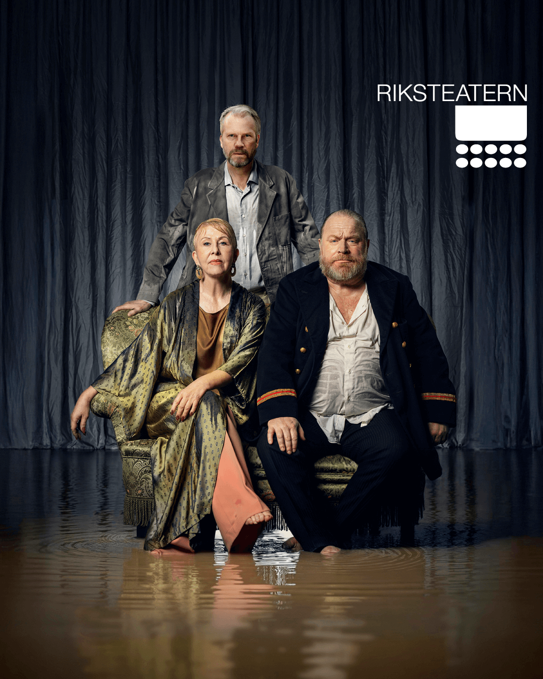 Tre personer sitter och står framför en mörk, draperad bakgrund. En kvinna sitter i mitten i en mönstrad klänning, en man i mörk kavaj sitter bredvid henne, och en tredje man står bakom dem. Alla ser allvarliga ut.