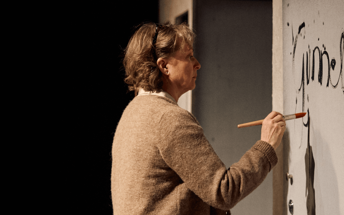 En person med ljust hår och glasögon, klädd i en beige tröja, står i profil och målar med pensel på en ljus vägg. På väggen syns svart text och former. Bakgrunden är mörk.