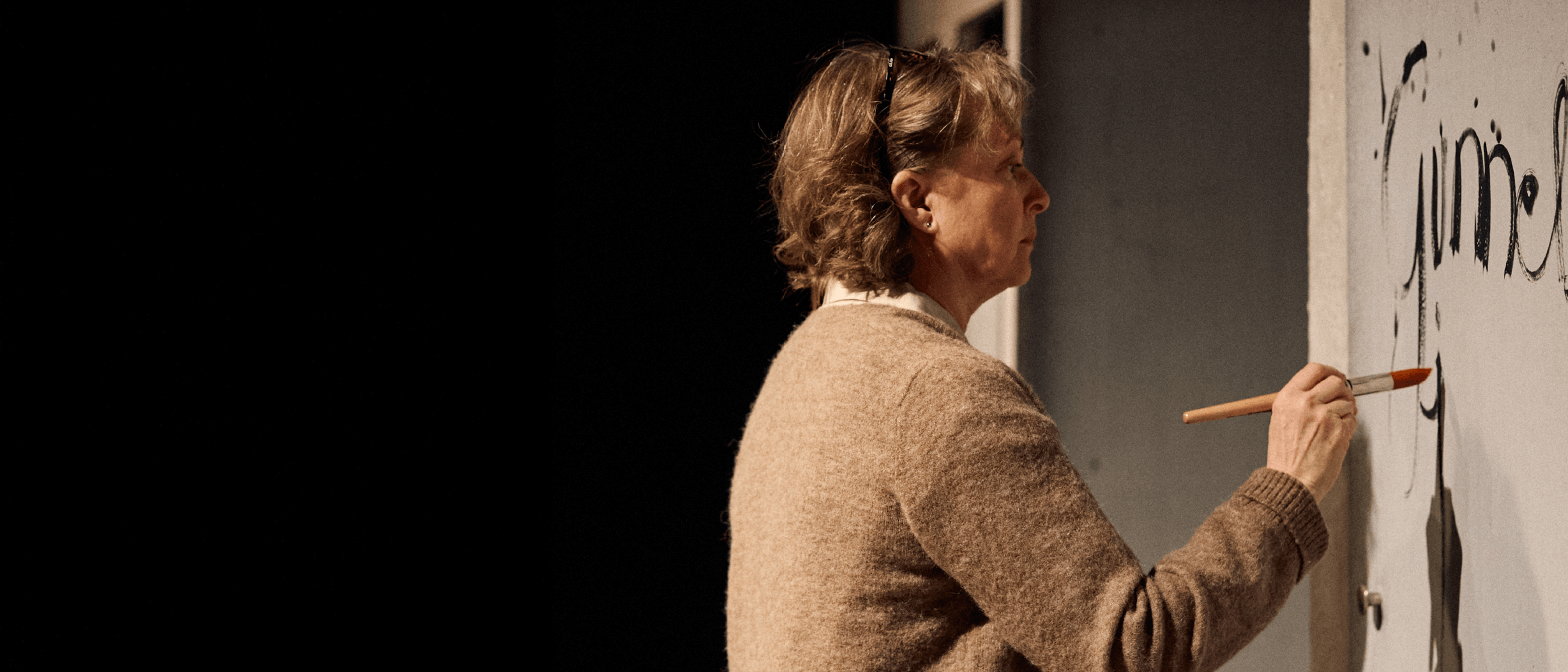 Vaaleahiuksinen ja silmälasipäinen henkilö, pukeutunut beigeen neuleeseen, seisoo sivuttain ja maalaa pensselillä vaaleaa seinää. Seinällä näkyy mustaa tekstiä ja kuvioita. Tausta on tumma.