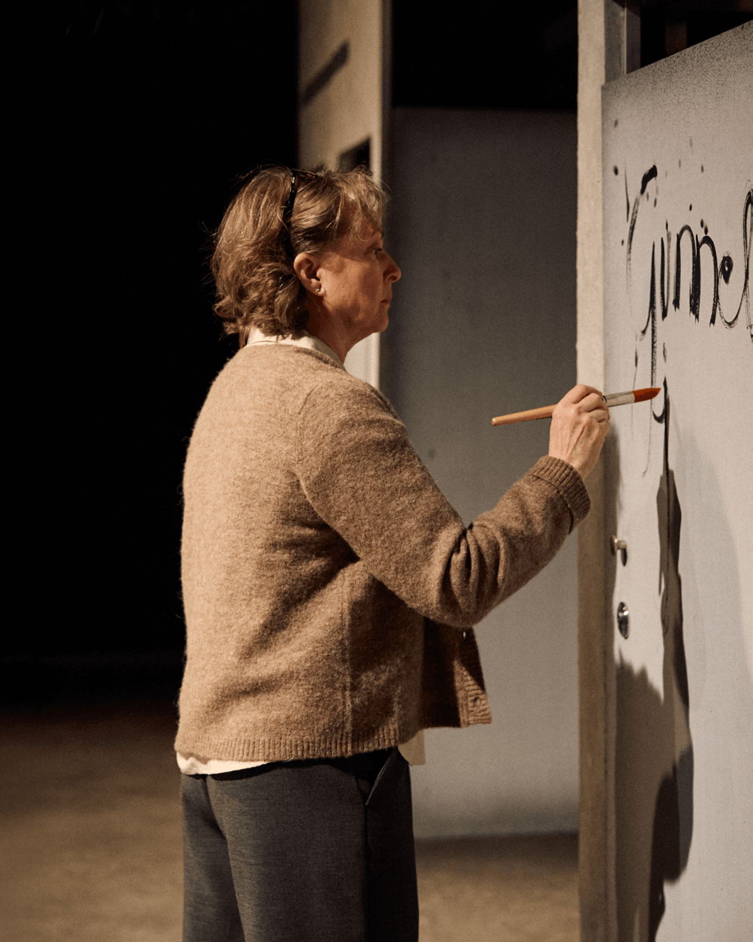 Vaaleahiuksinen ja silmälasipäinen henkilö, pukeutunut beigeen neuleeseen, seisoo sivuttain ja maalaa pensselillä vaaleaa seinää. Seinällä näkyy mustaa tekstiä ja kuvioita. Tausta on tumma.