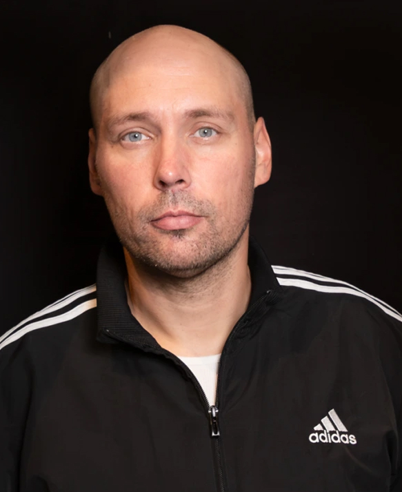 En person klädd i en svart Adidas-jacka med vita ränder på axlarna står mot en helt svart bakgrund. Jackan är uppknäppt upptill och under syns en vit tröja. Ljuset är jämnt och fokuserat på överkroppen, vilket ger en tydlig kontrast mot den mörka bakgrunden.