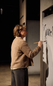 Henkilö maalaa tekstiä harmaalle seinälle siveltimellä.