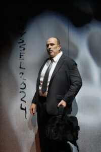 Henkilö puvussa seisoo mustan laukun kanssa seinän edessä, jossa lukee ”BRYSSEL 2024”.