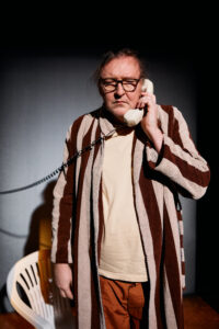 Henkilö seisoo raidallisessa aamutakissa ja puhuu vanhanaikaiseen lankapuhelimeen.