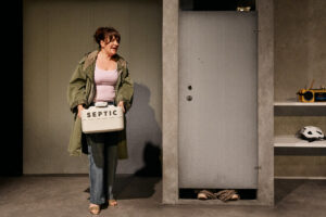 Henkilö pitelee valkoista säiliötä, jossa lukee ”SEPTIC”, harmaan oven edessä, hyllyn vieressä, jossa on radio ja kypärä.