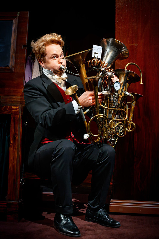 Dennis Nylund i frackliknande scenkostym sitter på en stol och spelar på ett stort, fantasifullt blåsinstrument sammansatt av flera trumpeter och horn. Scenen är mörk och träpaneler omger personen, medan instrumentets mässingsdetaljer glänser i ljuset.