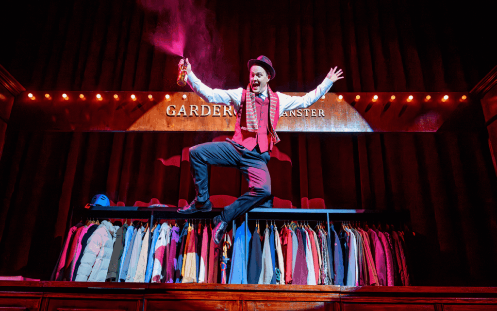 Skådespelaren Dennis Nylund som karaktären Garderobiären från föreställningen Kulturbärarna skuttar upp på en garderobsmöbel och slår klackarna ihop på en färgstark teaterscen.