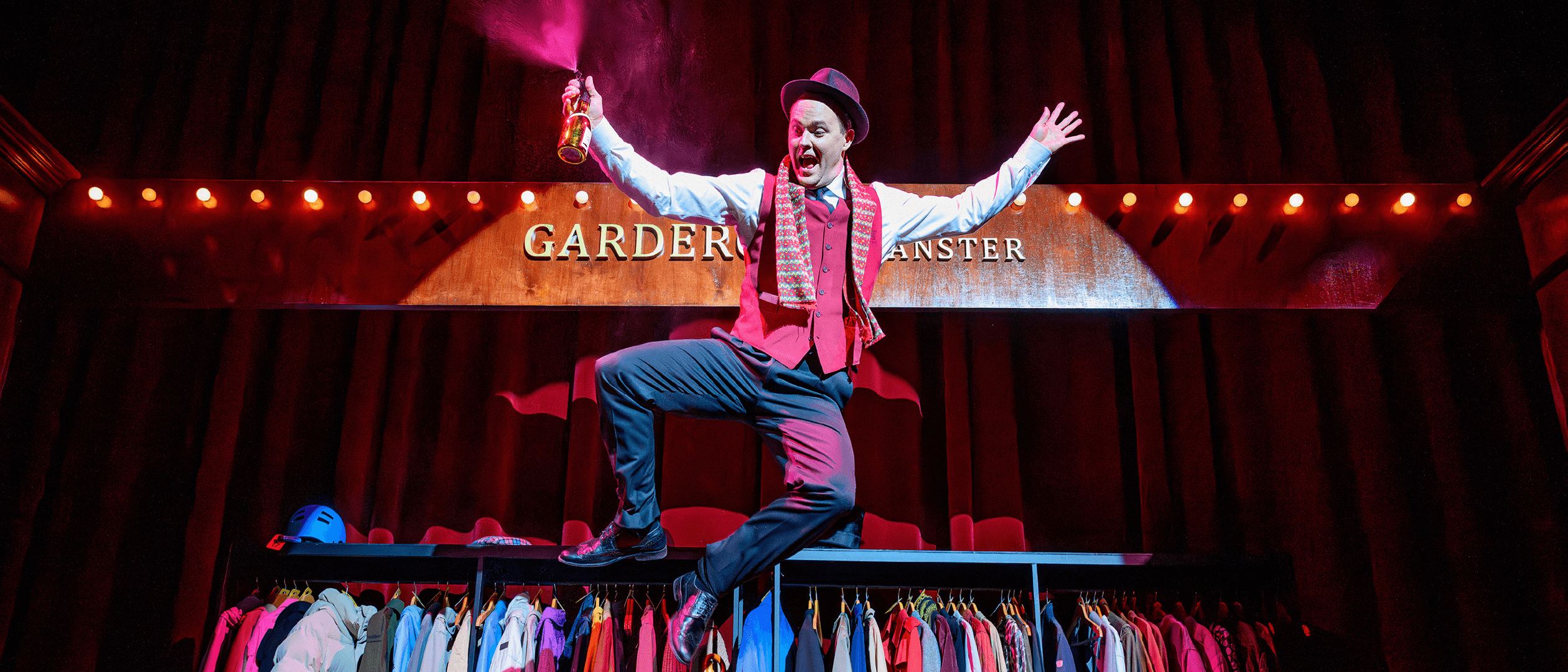 Skådespelaren Dennis Nylund som karaktären Garderobiären från föreställningen Kulturbärarna skuttar upp på en garderobsmöbel och slår klackarna ihop på en färgstark teaterscen.