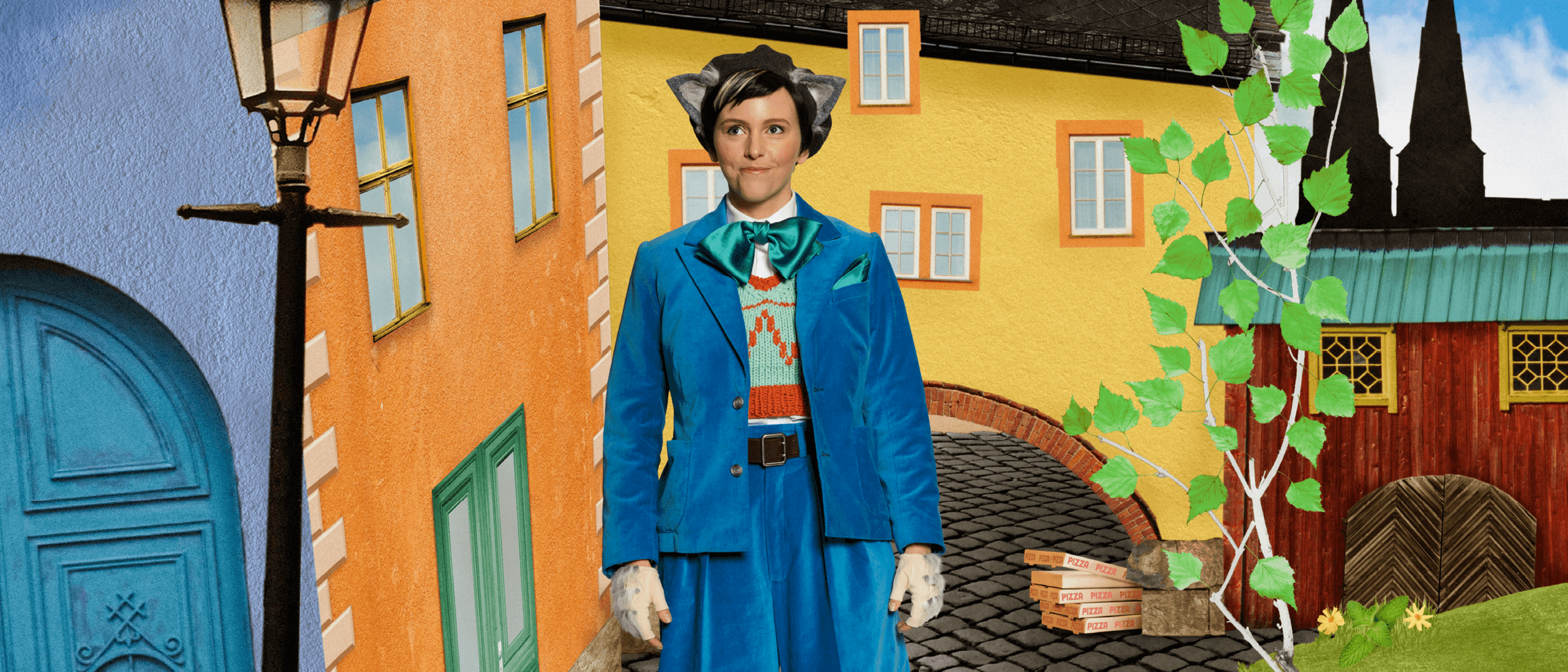 Skådespelaren Sonia Haga som Pelle Svanslös, klädd i blå kostym står på en kullerstensgata i ett fiktivt Uppsala, bland färgglada hus och blommor.