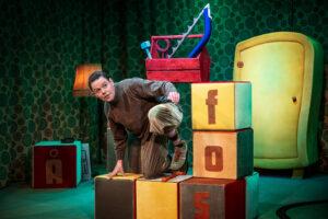 n scenbild med stora färggranna klossar med bokstäver, där en person klättrar över dem i ett lekfullt rum med mönstrade väggar och barnrumsrekvisita.