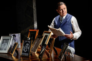 Skådespelaren Patrick Henriksen står framför en rad inramade porträtt som placerats på en flyger. han föreställer Amos Anderson och håller en tidning i handen