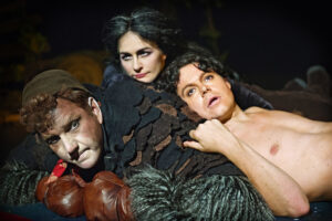 En teaterscen i Djungelboken där tre personer ligger tätt intill varandra; en person i pälsliknande kostym, en annan bar överkropp och en tredje bakom dem, allt belyst i varmt scenljus.