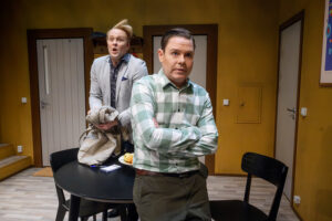 En scen i ett hemmiljö‑liknande rum där två personer står vid ett runt bord; den ena med armarna i kors och den andra hållande en jacka, i en vardaglig och lätt dramatisk situation.