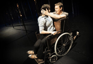 En kvinnlig skådespelare sitter i knät på Patrick Henriksen som sitte i en rullstol. Han håller med båda händerna om kvinnans huvud och talar till henne.