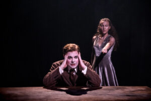 En teaterscen där en person sitter vid ett bord med händerna mot huvudet, medan en annan person står bakom och sträcker ut en arm mot honom i ett dramatiskt ljus.