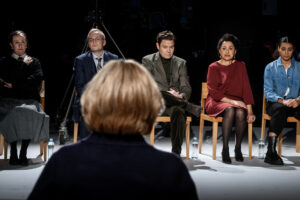 En teaterscen där en person sitter vänd mot en grupp människor som sitter på stolar i en halvcirkel, som om de deltar i ett samtal, en utfrågning eller ett möte i en dramatisk scen.