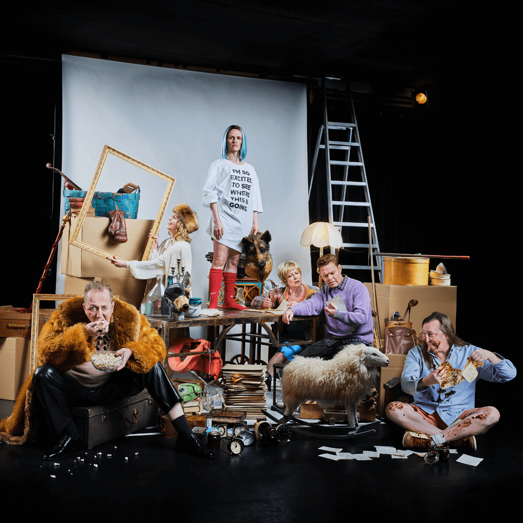 The actors in the play Nattland standing in a theatrical scene, surrounded by props, boxes, and furniture.