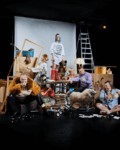 En teatral och humoristisk gruppscen där flera personer sitter och står bland en mängd rekvisita – kartonger, ramar, kläder, stege, böcker och ett gosedjur – uppställda som ett överlastat stilleben mitt på scenen.