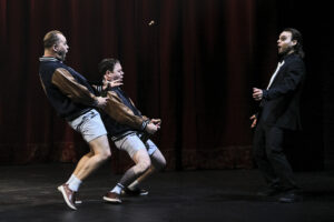 Tre personer på en teaterscen, två i sportiga jackor och shorts som gör en fysisk, nästan slapstick‑lik rörelse, medan en person i kostym står mittemot dem framför en mörkröd ridå.