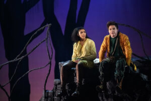 Två personer sitter på en klippkant i skogen på en teaterscen, omgivna av mörka trädgrenar mot en lilatonad himmel, i en stämningsfull och äventyrlig miljö.