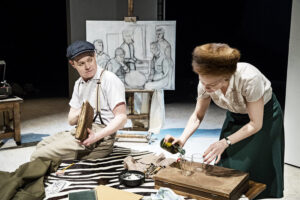 Patrick sitter på golvet ikklädd vit skjorta med uppkavlade ärmar. Han sitter med Tove Jansson som målar på golvet. Hon häller färg i ett glas omgivet av penslar och papper att måla på . Tove har pälsmössa på sig.