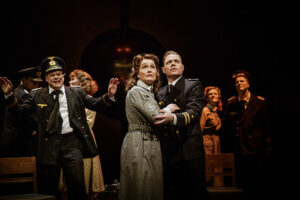 n teaterscen där personer i uniformer och tidsenliga kläder står samlade, med två figurer i centrum som håller om varandra, belysta av varmt scenljus mot en mörk bakgrund.