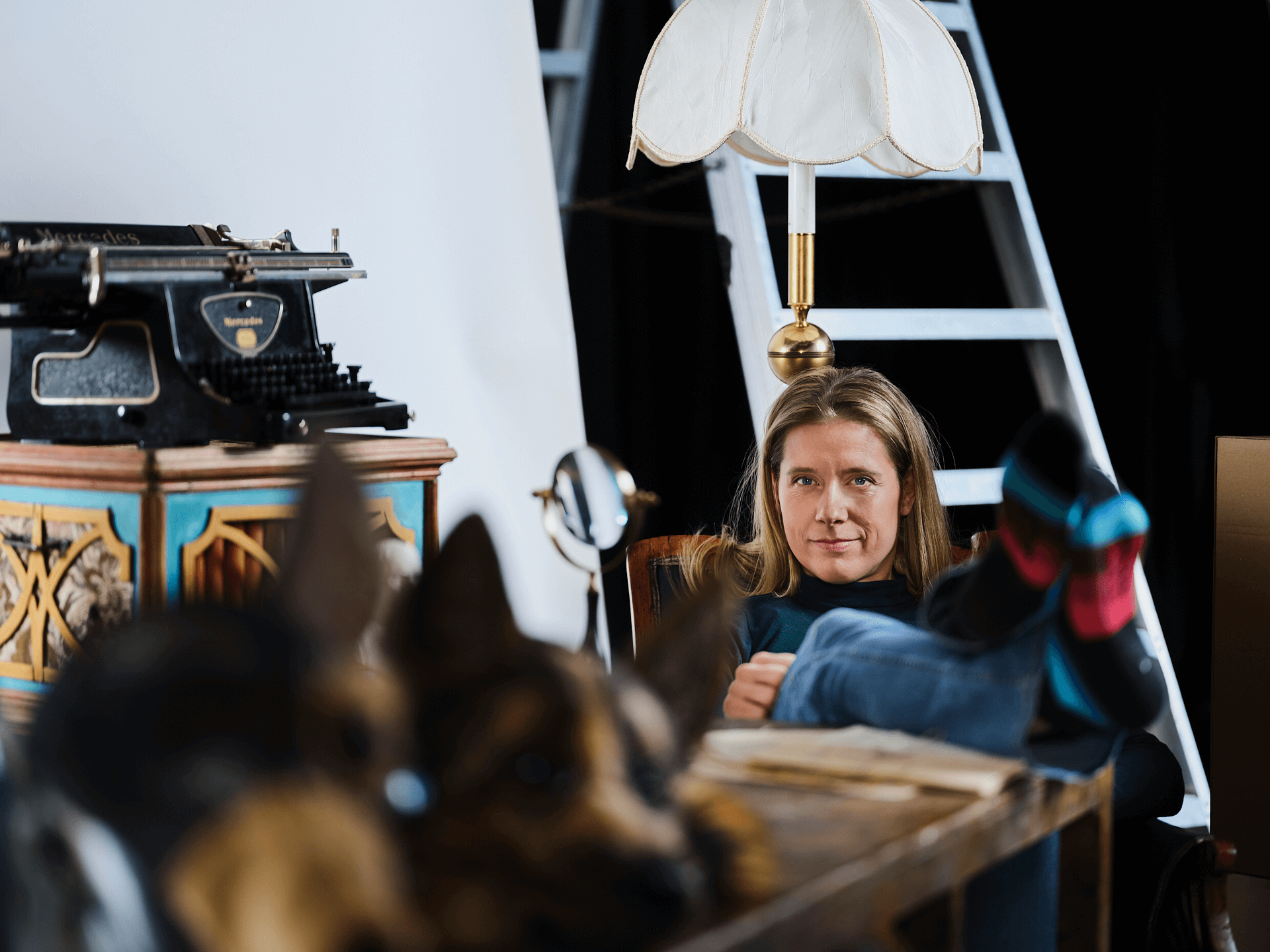 Anna-Maria Lipponen, regissör sitter avslappnat med fötterna upp på ett bord i ett rum fyllt av teaterrekvisita, inklusive en gammaldags skrivmaskin, en dekorativ trälåda, en bordslampa och en stege i bakgrunden.