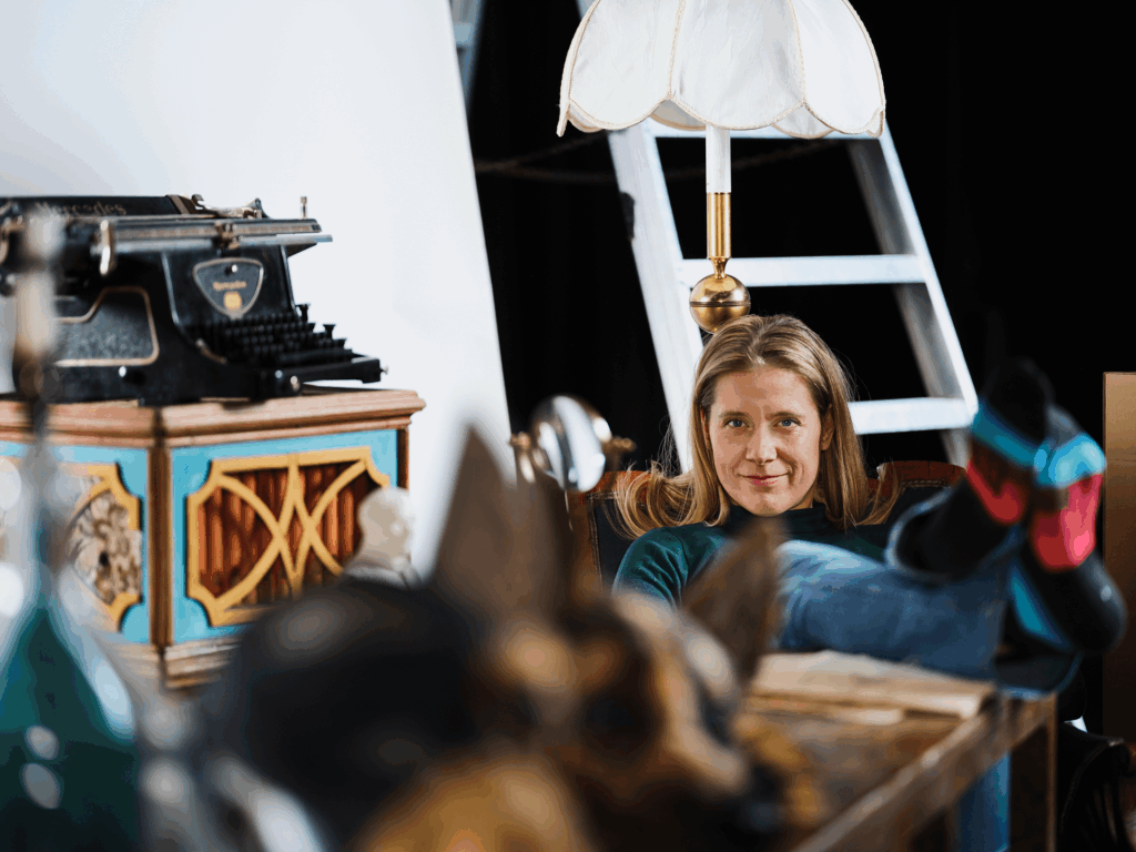 En person sitter avslappnat med fötterna upp på ett bord i ett rum fyllt av teaterrekvisita, inklusive en gammaldags skrivmaskin, en dekorativ trälåda, en bordslampa och en stege i bakgrunden.