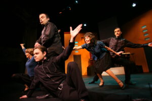 En grupp personer utför en dynamisk och uttrycksfull koreografi på en teaterscen, med stora rörelser och utspridda kroppar framför en färgstark scenografi.