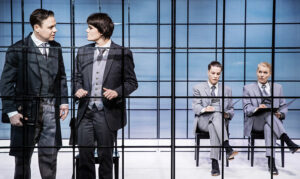 Skådespelarna Patrick Henriksen och Joanna Haarti står framför en glasvägg i blått sken med svarta rutor. Bakom denna vägg sitter två män som följer med vad som händer