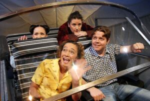 Fyra personer sitter tillsammans i en öppen vagn eller åkattraktion med rutiga säten, i en scen som förmedlar rörelse och vardaglig gemenskap.