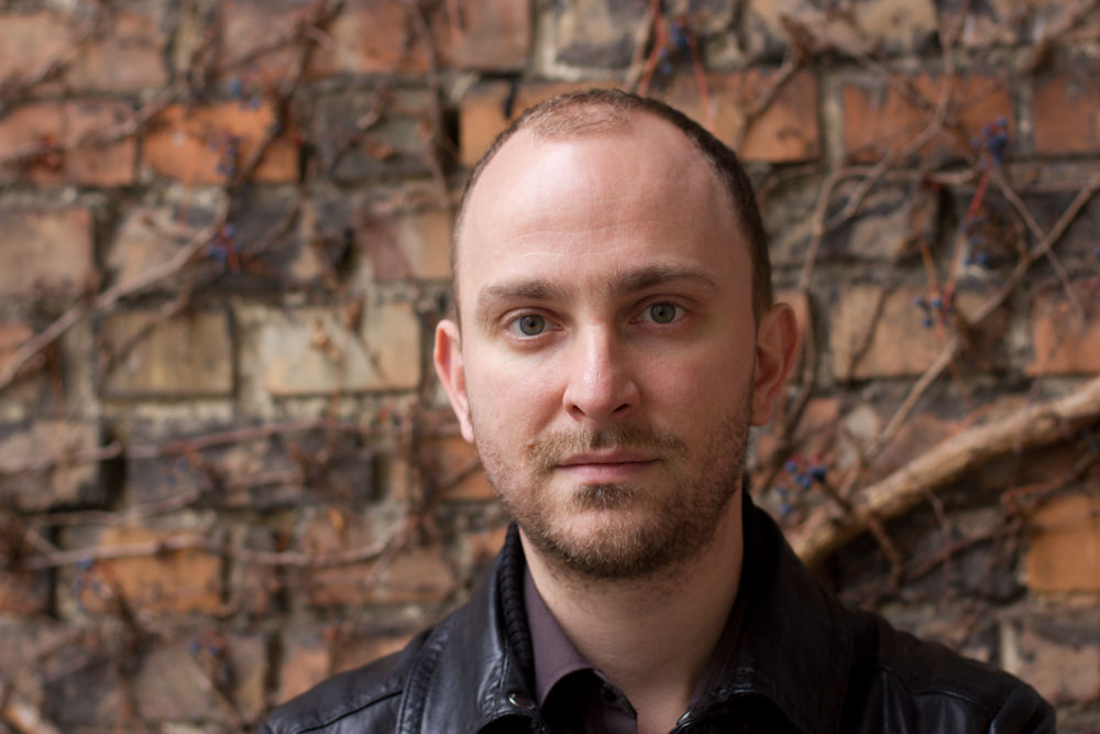 Författaren till Nattland, Marius von Mayenburg framför en rostbrun tegelvägg med vissnande klängväxt utan blad i bakgrunden