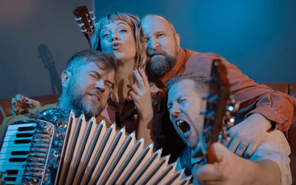 Artisterna i föreställningen Ballader och oförskämdheter, med Paleface i ensemblen, sitter tätt tillsammans med dragspel och gitarrer i en lekfull gruppbild under blå scenbelysning.