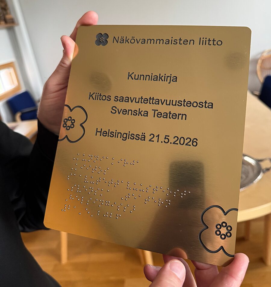 En person håller upp en guldplakett med ingraverad text. Plaketten är utdelad av Näkövammaisten liitto och innehåller texten ”Kunniakirja – Kiitos saavutettavuusteosta Svenska Teatern – Helsingissä 21.5.2026”. Nederst finns punktskrift.