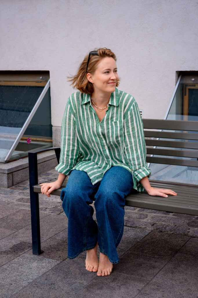 Sanna Nedergård sitter barfota på en bänk utomhus framför en ljus byggnadsfasad. Personen bär en grön- och vitrandig skjorta, vida jeans och solglasögon på huvudet, och är vänd åt sidan. I bakgrunden syns markbeläggning av sten samt låga fönster med öppna glasluckor.